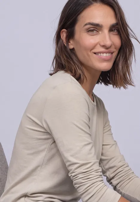 Une femme souriante aux cheveux bruns mi-longs, vêtue d'un haut à manches longues beige sur fond clair.