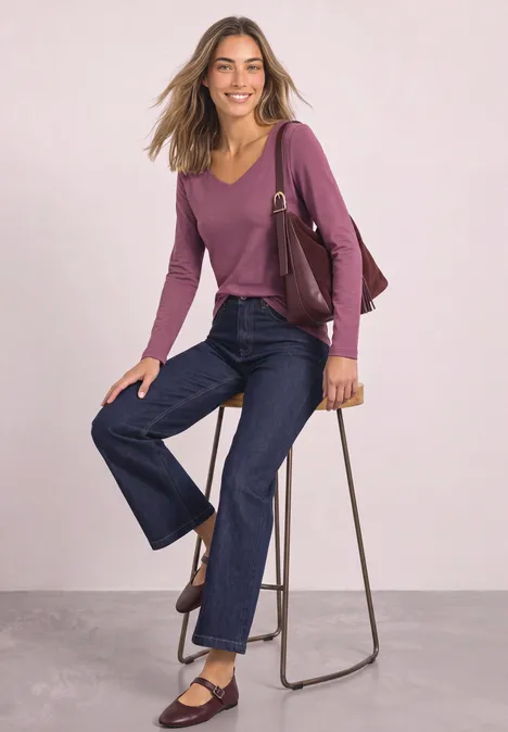 Femme assise sur tabouret, pull violet, jean large retroussé, chaussures et sac marron.