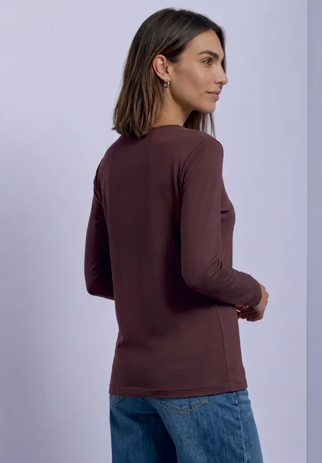 Femme de profil portant un haut marron à manches longues et un jean sur fond clair.