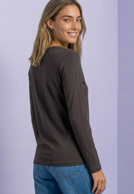 Une femme souriante portant un t-shirt à manches longues noir et un jean bleu devant un fond clair.