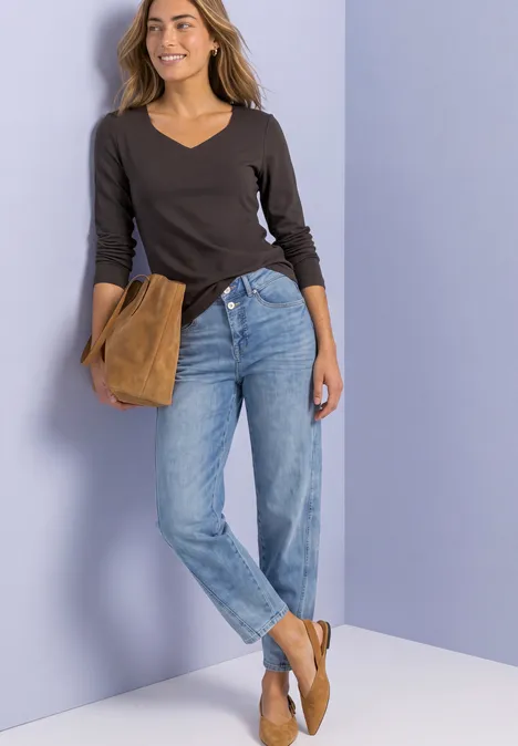 Femme en pull à col V foncé, jean bleu et chaussures marron, tenant une pochette camel devant un mur violet pastel.
