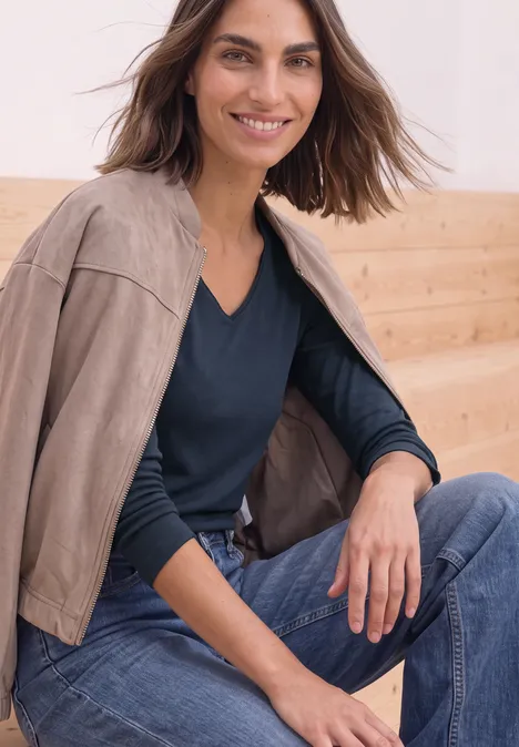 Vrouw zit, suède jas los over schouders, donkerblauwe trui en jeans, lacht voor houten wand.