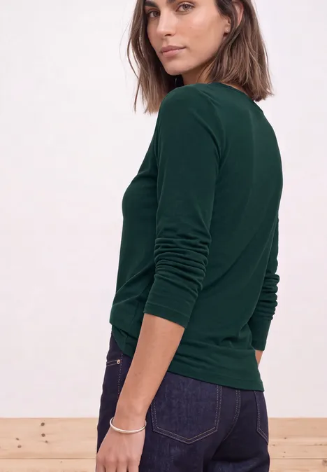 Frau dreht sich leicht zur Kamera, trägt grünes Langarmshirt und dunkle Jeans