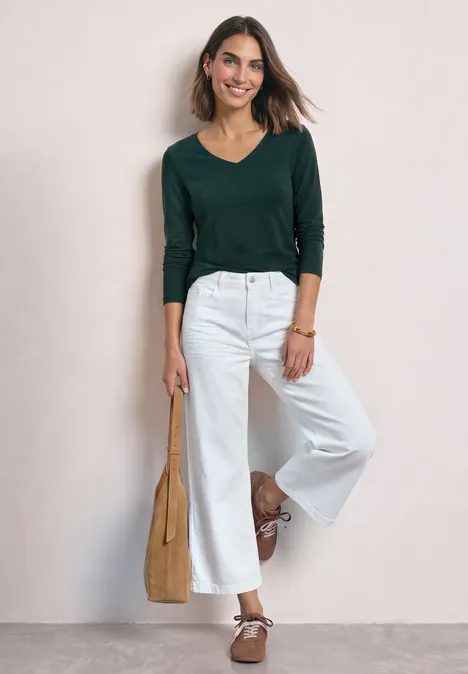 Femme portant un haut vert à manches longues et un pantalon blanc ample, avec des ballerines marron et un sac beige.