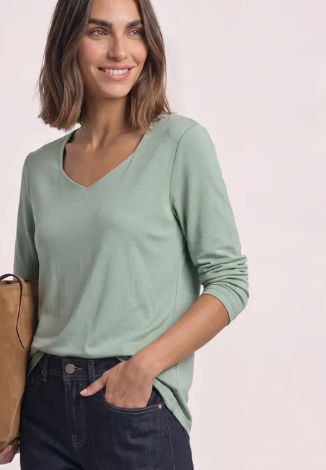 Une femme souriante porte un haut vert à manches longues avec col en V et un jean foncé