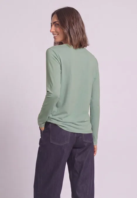 Femme en haut vert à manches longues, de profil, main dans la poche et regard souriant par-dessus l'épaule.