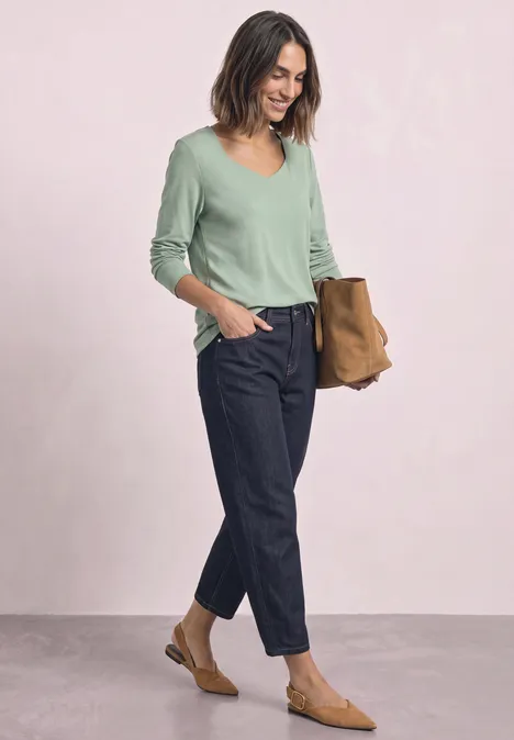 Femme en pull vert col V, pantalon bleu marine et mocassins bruns tenant un grand sac.