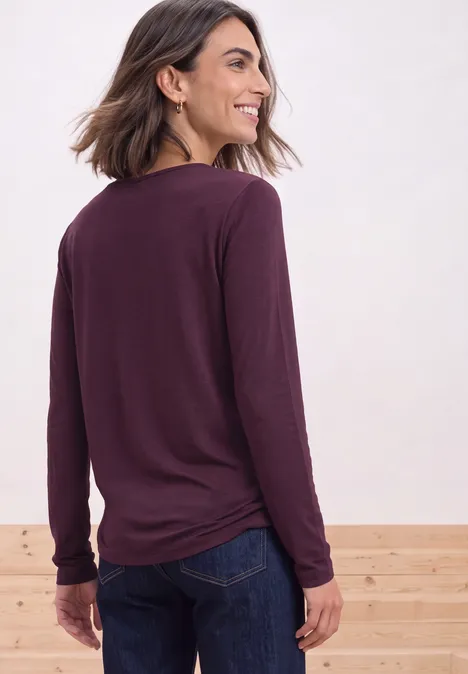 Vrouw van achteren in bordeauxrood shirt met lange mouwen en spijkerbroek, glimlachend over haar schouder.