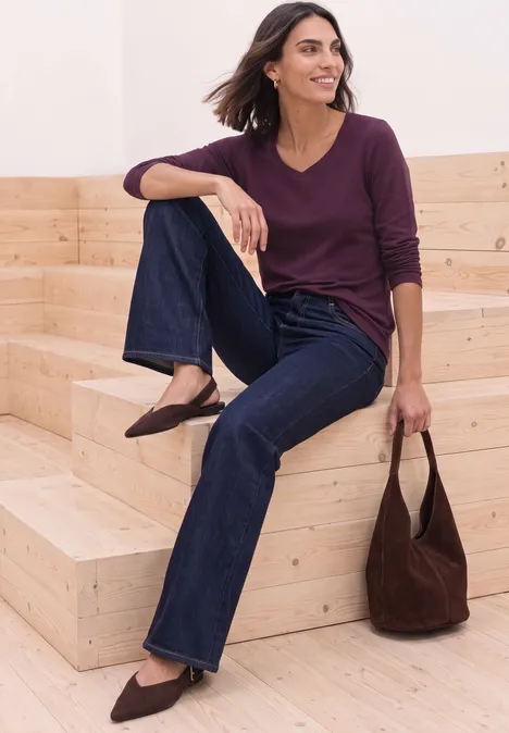 Vrouw zit relaxed op houten treden, draagt jeans, donkerpaarse longsleeve en spitse schoenen, houdt een schoudertas vast.