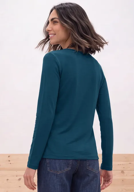 Vrouw van achteren in petrolkleurige longsleeve en spijkerbroek, halflang bruin haar, licht zijwaarts kijkend.