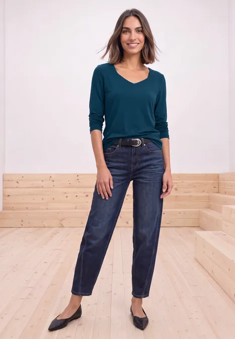 Vrouw in petrolkleurig shirt, donkere jeans, riem en zwarte ballerina’s op licht houten treden.