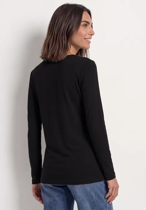 Frau mit schulterlangem dunklem Haar in schwarzem Langarmshirt und blauen Jeans steht seitlich vor weißer Wand.