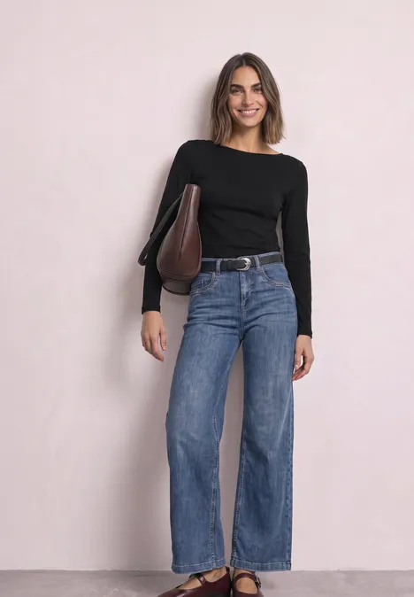 Junge Frau steht vor hellem Hintergrund, trägt schwarzen Pullover, weite Jeans und lächelt.