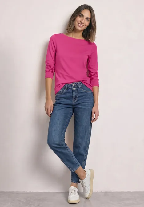 Langarm Shirt mit U-Boot-Ausschnitt magenta dream Frau lehnt lässig an Wand, trägt pinken Pullover, blaue Jeans und weiße Sneaker.