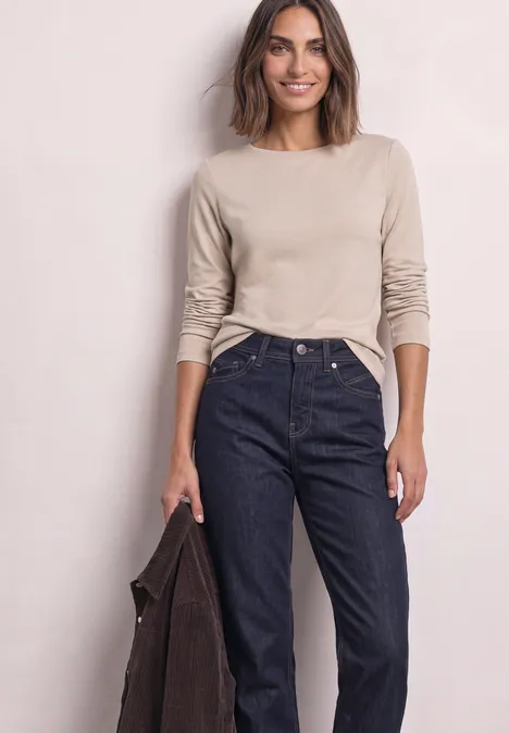 Frau mit beigeem Langarmshirt und hoher Jeans, hält dunklen Mantel in einer Hand, lächelt vor hellem Hintergrund.