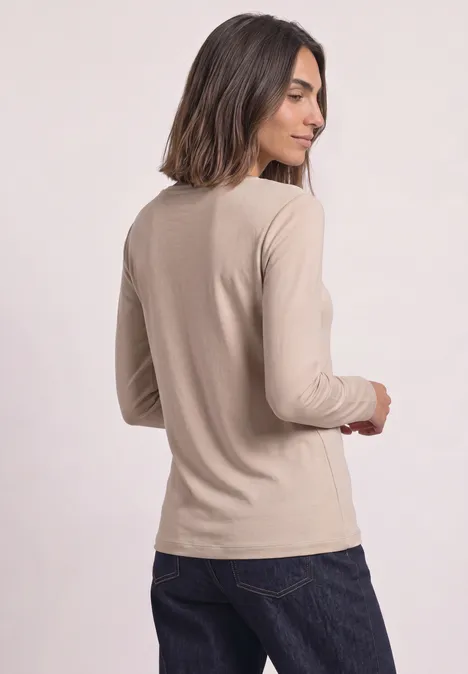 Frau mit schulterlangen dunklen Haaren trägt beige Langarmshirt und dunkle Jeans vor hellem Hintergrund.