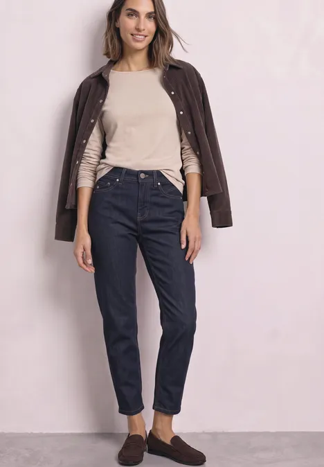 Frau mit beiger Bluse, dunkler Lederjacke über den Schultern, blauen Jeans und braunen Schuhen vor rosafarbenem Hintergrund.
