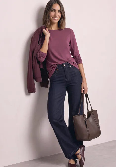 Eine lächelnde Frau in lila Shirt und Jeans lehnt an der Wand, hält eine braune Tasche.