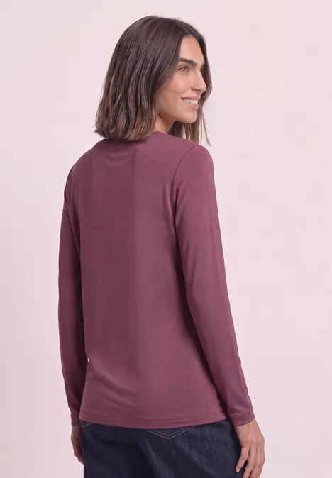 Frau mit schulterlangem Haar in lässigem langärmligem Shirt in Mauve, schaut über die Schulter und lächelt.