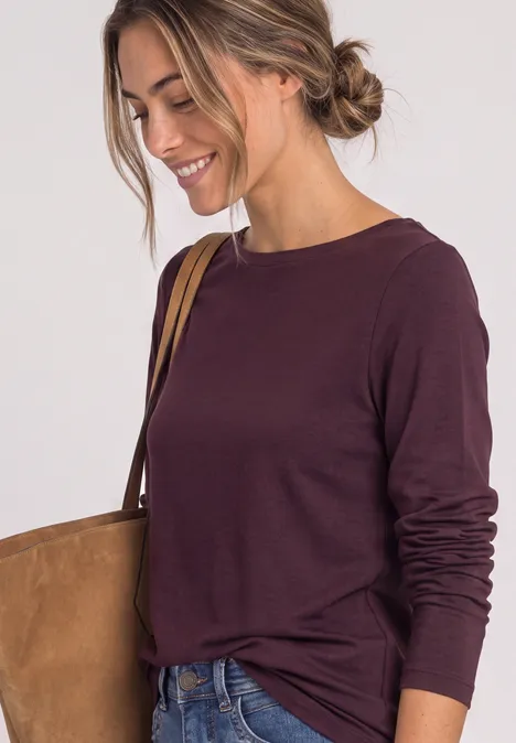 Frau in kastanienfarbenem Langarmshirt und Jeans mit Umhängetasche lächelnd nach unten blickend.