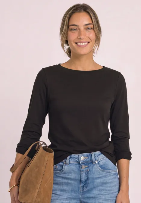 Eine lächelnde Frau in schwarzem Langarmshirt und Jeans hält eine braune Wildledertasche vor hellem Hintergrund.