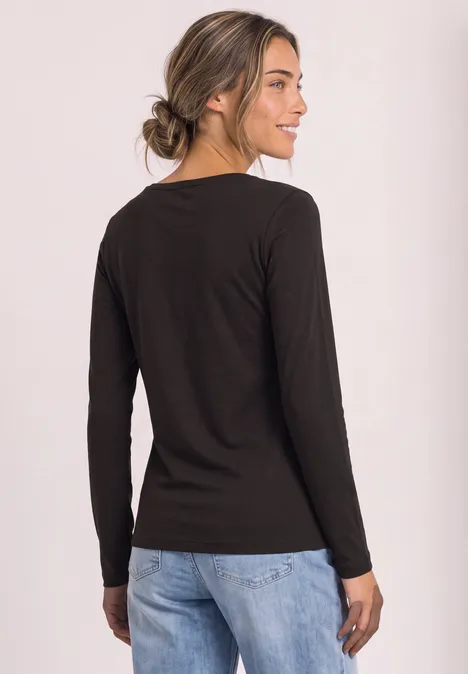 Eine Frau in schwarzem Langarmshirt und blauen Jeans steht leicht seitlich, lächelnd.