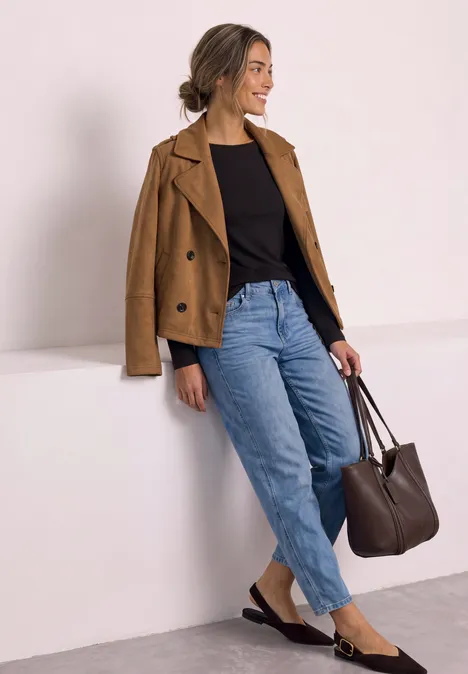 Eine Frau in lässiger Kleidung mit brauner Jacke, schwarzem Shirt, Jeans und Tasche lehnt an einer weißen Wand.
