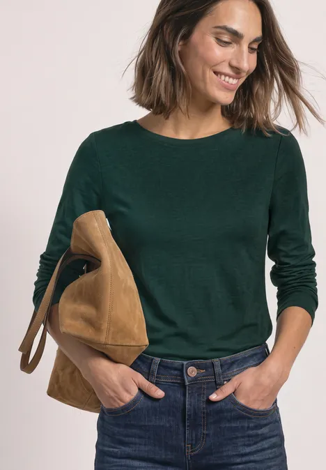 Vrouw lacht, draagt een donkergroene trui, spijkerbroek en een bruine suède tas over haar schouder.