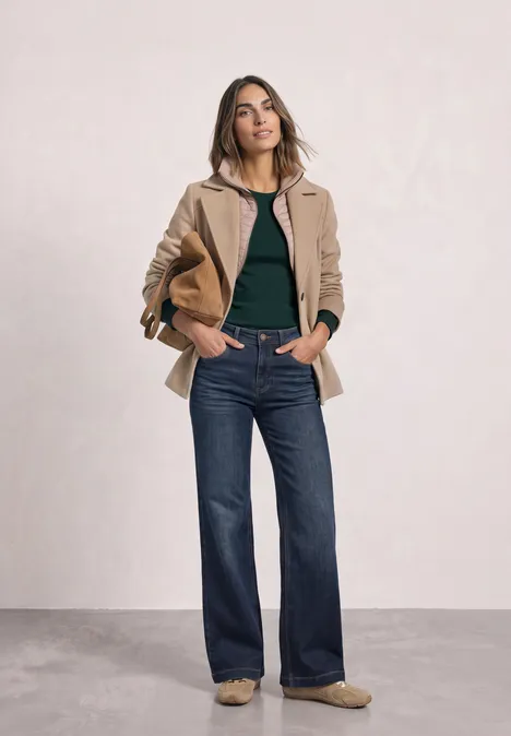 Een vrouw draagt blauwe wijde jeans, een zwarte top en een beige jas, met een papieren tas in haar hand.