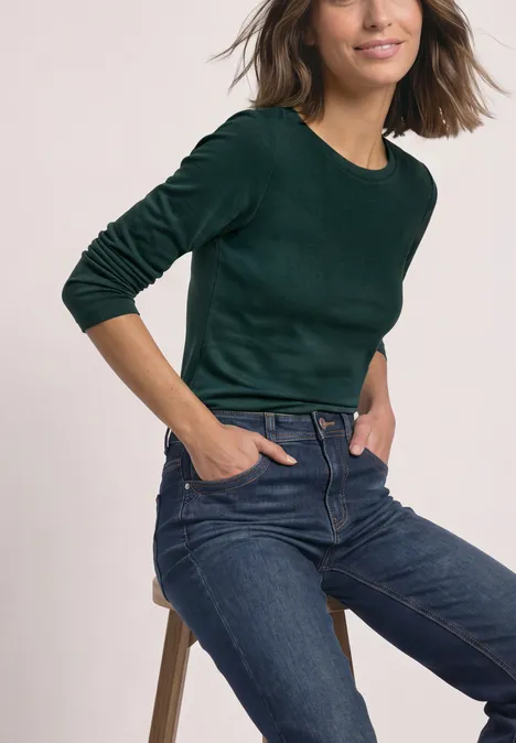 Vrouw zit op een kruk, draagt een groen shirt met lange mouwen en een blauwe spijkerbroek, handen in zakken.