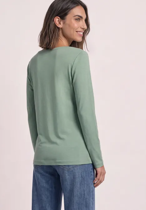 Eine Frau steht seitlich abgewandt, trägt grünes Langarmshirt und blaue Jeans und lächelt.