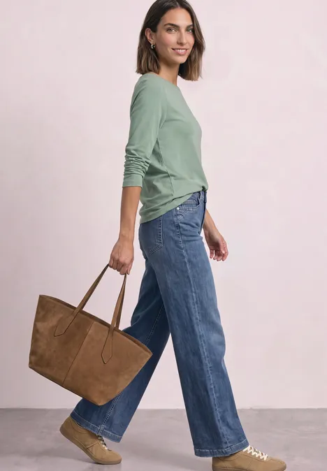 Frau trägt grünes Oberteil, weite Jeans, braune Tasche und Sandalen vor hellem Hintergrund.