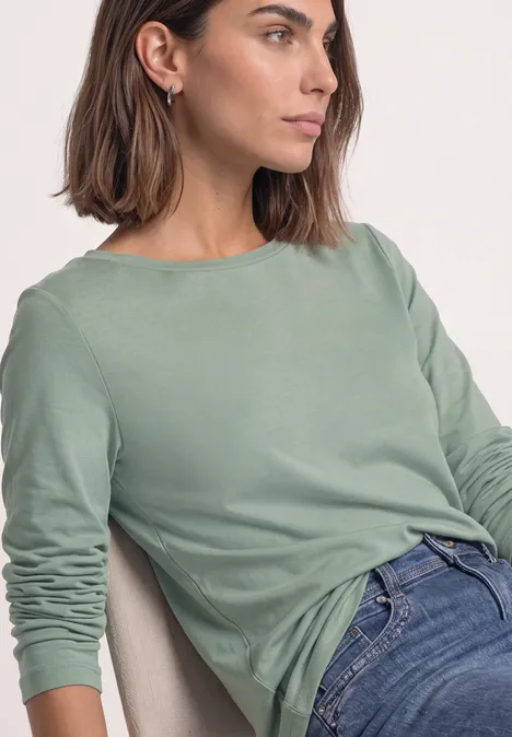 Seitlich abgebildete Frau in legerem, mintgrünem Langarmshirt und Jeans, die auf einem Stuhl sitzt.