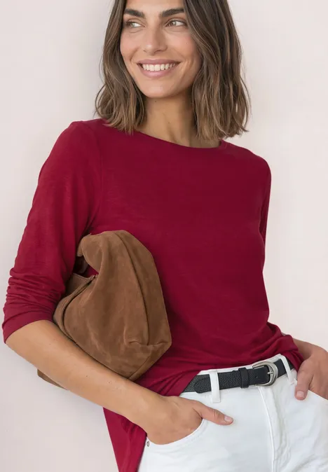Frau in rotem Langarmshirt, weißer Hose und schwarzem Gürtel, lächelnd mit brauner Tasche.