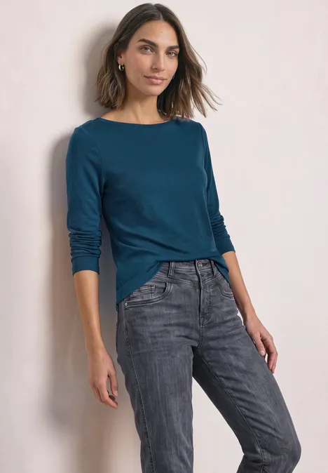 Vrouw in blauwgroene trui met lange mouwen en grijze jeans, glimlachend voor een neutrale achtergrond.