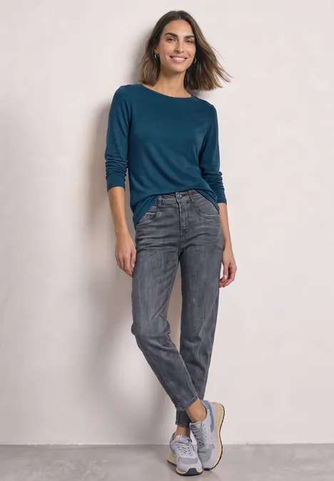 Een vrouw draagt een blauwgroen shirt, grijze jeans en witte sneakers tegen een lichte achtergrond.