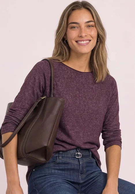 Frau in lässigem lila Pullover und Jeans, trägt eine braune Tasche und lächelt.