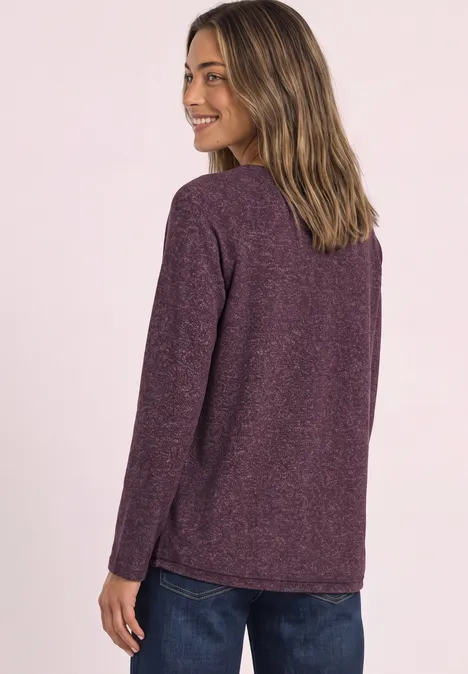 Rückansicht einer Frau im violetten Pullover, sie lächelt über die Schulter vor hellem Hintergrund.