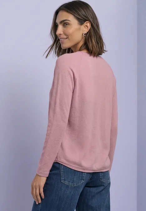 Frau mit rosa Pullover und blauer Jeans blickt über die Schulter vor hellem Hintergrund.