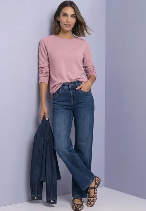 Frau im rosa Pullover, blauen Jeans und Leoparden-Sandalen hält eine Jeansjacke vor lila Hintergrund.