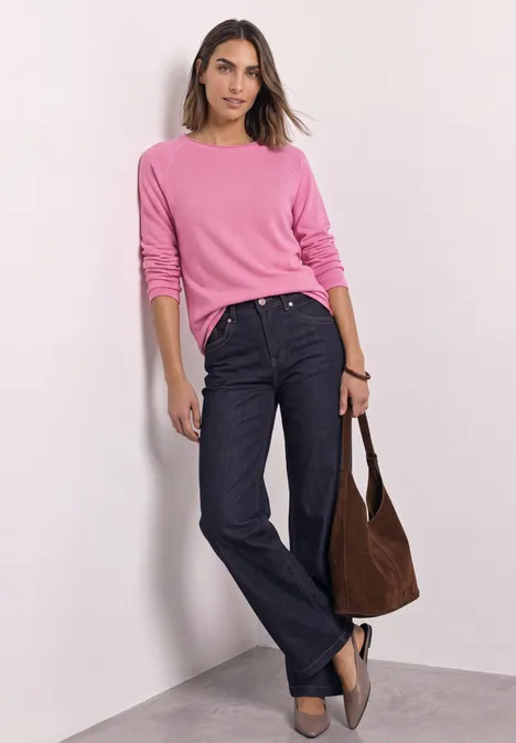 Langarm Shirt mit Rundhals und Rollkante warm blush mel. Eine Frau lehnt lässig an einer Ecke, trägt ein pinkes Oberteil, dunkelblaue Jeans und hält eine braune Tasche.