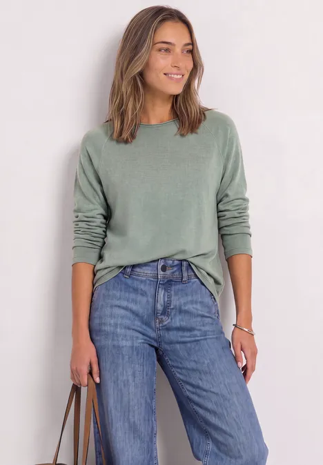 Langarm Shirt mit Rundhals und Rollkante eucalyptus green mel. Eine lächelnde Frau in grünem Pullover und blauen Jeans hält eine Tasche.