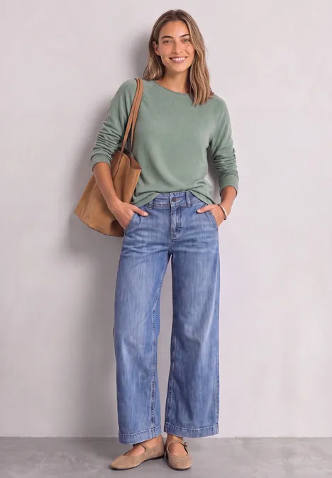 Langarm Shirt mit Rundhals und Rollkante eucalyptus green mel. Junge Frau steht lächelnd vor heller Wand, trägt grünen Pullover, weite Jeans, braune Tasche und Loafers.