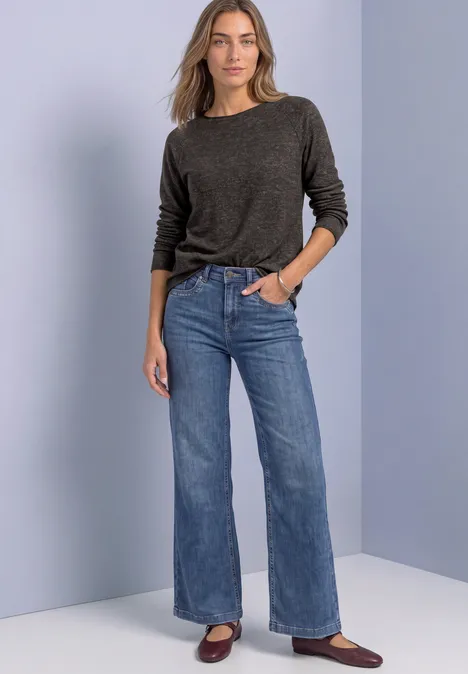 Vrouw in grijze trui, high-waist wijde jeans en bruine schoenen tegen een neutrale achtergrond