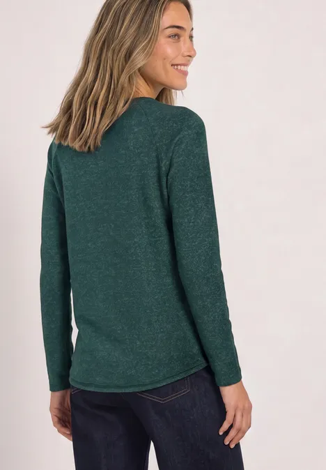 Vrouw van achteren in een groen shirt met lange mouwen en donkere spijkerbroek, ze lacht opzij.