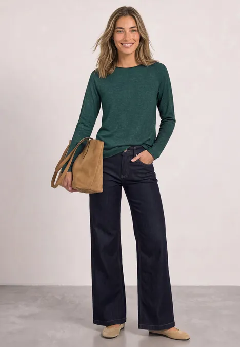 Vrouw lachtend in groen shirt, wijde donkerblauwe broek en beige tas voor neutrale achtergrond.