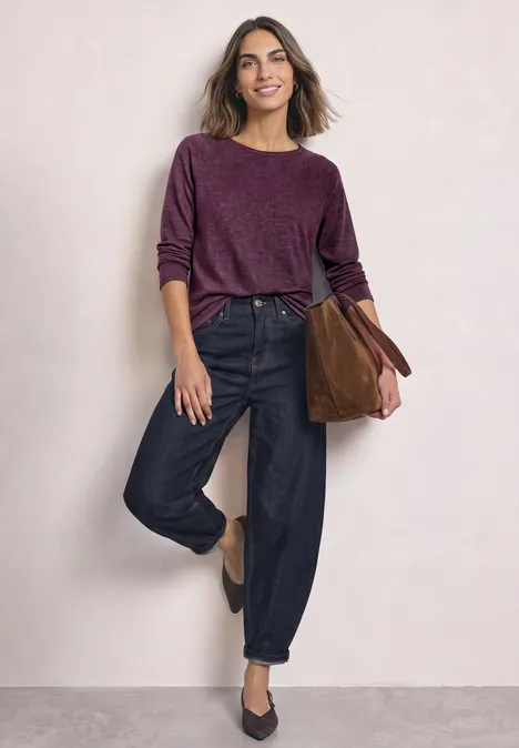 Een lachende vrouw draagt een bordeauxrode trui, zwarte jeans en heeft een bruine tas.