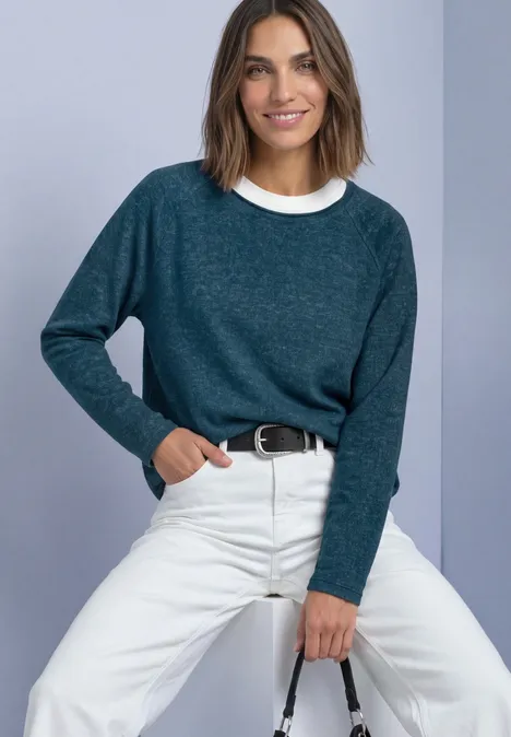 Vrouw zittend voor lichte achtergrond, blauwgroene trui, witte broek en glimlach.