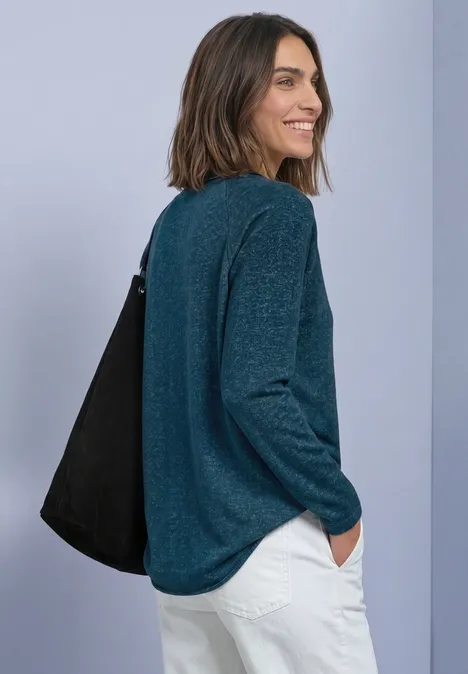Vrouw in groene trui en witte broek, glimlachend over haar schouder kijkend met een zwarte tas.