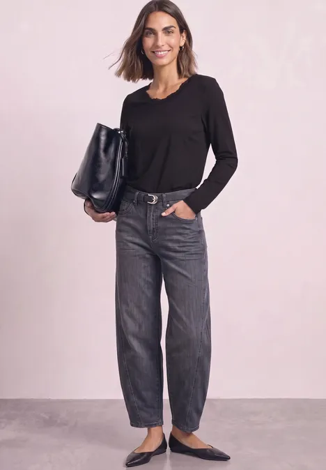 Frau in schwarzem Langarmshirt, weiten Jeans und schwarzen Ballerinas mit großer schwarzer Tasche vor hellem Hintergrund